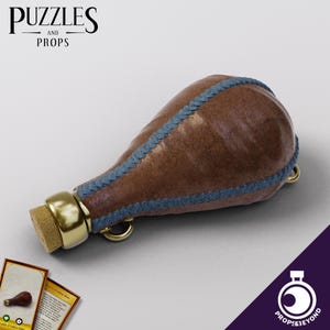 Peut inclure: Une fiole en cuir marron avec des coutures bleues et un bouchon en métal doré. La fiole a un bouchon en liège et une boucle en métal.