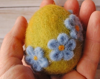 May include: A yellow felt Easter egg decorated with blue felt flowers. The egg is being held in a hand.