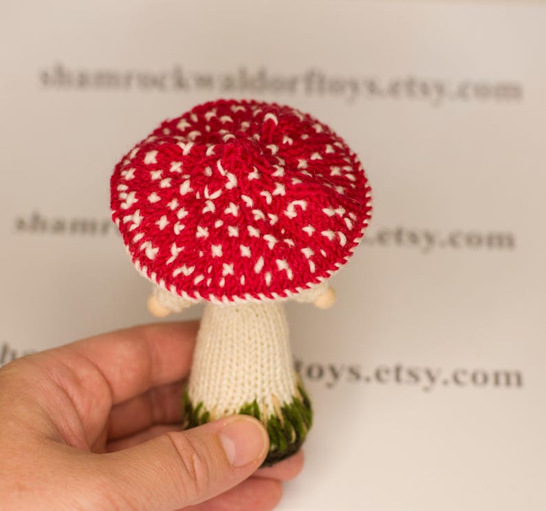 May include: A hand is holding a knitted mushroom toy with a red and white spotted cap and a white stem. The mushroom is approximately 15cm tall.