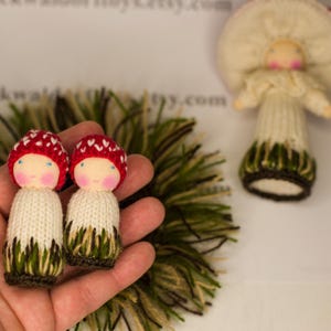 May include: Two small knitted dolls with red mushroom caps and white bodies. The dolls are wearing green and brown knitted skirts. The dolls are being held in a hand.
