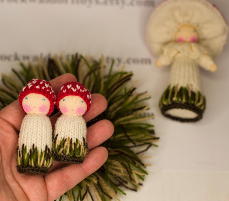 May include: Two small knitted dolls with red mushroom caps and white bodies. The dolls are wearing green and brown knitted skirts. The dolls are being held in a hand.