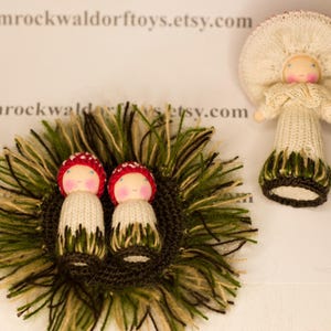 May include: Two knitted mushroom dolls with red caps and white bodies are nestled in a bed of green and brown yarn. The dolls have painted faces and are wearing green and brown skirts.