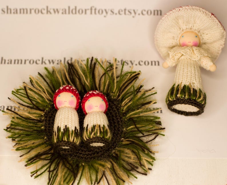 May include: Two knitted mushroom dolls with red caps and white bodies are nestled in a bed of green and brown yarn. The dolls have painted faces and are wearing green and brown skirts.