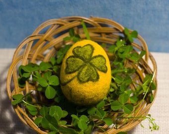 May include: A yellow, egg-shaped felt ornament with a green shamrock design, nestled in a woven basket with green clover leaves. The basket is made of light brown wicker and sits on a white surface with a blue background.