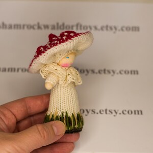 May include: A hand-knitted doll wearing a white dress with a lace collar and a red and white mushroom hat. The doll has a wooden head with painted blue eyes.