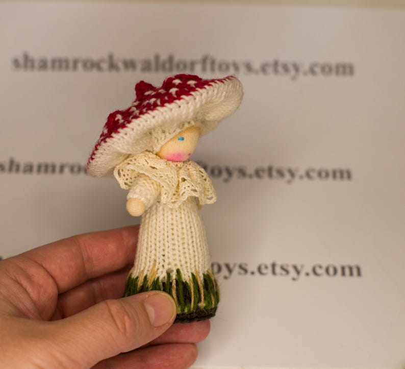 May include: A hand-knitted doll wearing a white dress with a lace collar and a red and white mushroom hat. The doll has a wooden head with painted blue eyes.