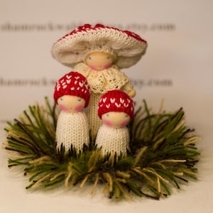 May include: Three knitted mushroom dolls with red and white hats. The dolls are sitting in a nest of green and brown yarn.