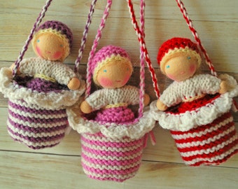 May include: Three handmade knitted dolls hanging from strings. Each doll is wearing a different colored dress and has a white crocheted basket. The dolls are pink, purple, and red.