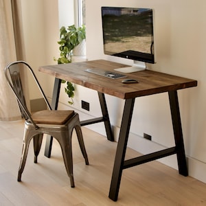 May include: A wooden desk with a dark finish and black metal legs, holding a computer monitor, keyboard, and mouse. A metal chair with a wooden seat is positioned beside the desk. The room has a window and a potted plant.