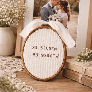 May include: A round, wooden embroidery hoop with a cream-colored, striped fabric. The text "35.5209°N -88.9306°W" is embroidered on the fabric. A white ribbon is attached to the top. A framed wedding photo is in the background.