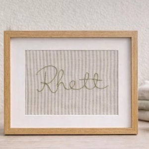 May include: A framed piece of art with the name "Rhett" embroidered in green thread on a striped fabric. The frame is light brown wood with a white mount. The artwork is a decorative item, suitable for a nursery or child's room.
