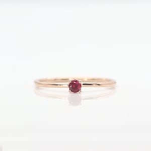 May include: A delicate rose gold ring with a single, round ruby gemstone set in a simple prong setting.