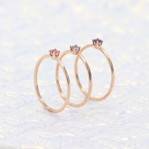 May include: Three delicate gold rings with small gemstones. The rings are stacked on top of each other. The gemstones are pink, purple, and a darker shade of purple.