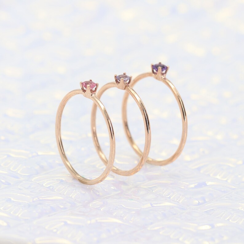 May include: Three delicate gold rings with small gemstones. The rings are stacked on top of each other. The gemstones are pink, purple, and a darker shade of purple.