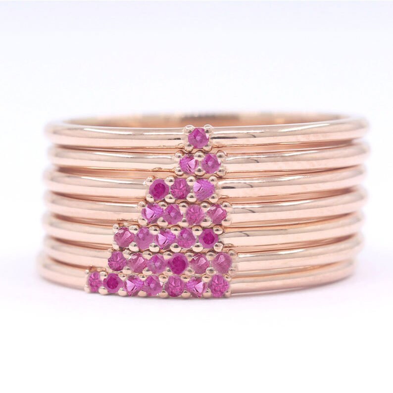 Puede incluir: Una pila de ocho anillos de oro rosa con un dise&ntilde;o de piedras preciosas rosas. Las piedras preciosas est&aacute;n dispuestas en forma de tri&aacute;ngulo en el anillo superior.