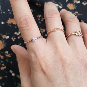 May include: Three gold rings on a finger. The ring on the index finger has a small purple gemstone. The ring on the middle finger is a simple band. The ring on the pinky finger has a small flower design.