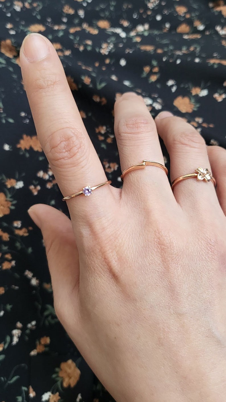 May include: Three gold rings on a finger. The ring on the index finger has a small purple gemstone. The ring on the middle finger is a simple band. The ring on the pinky finger has a small flower design.