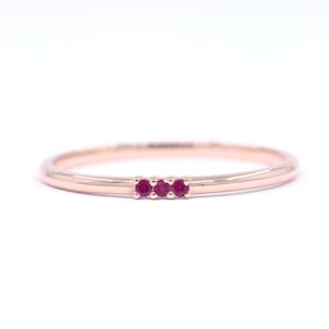 May include: A slender rose gold ring adorned with three small, vivid red gemstones. The ring has a thin band and a minimalist design. The gemstones are evenly spaced across the top of the ring. The ring is suitable for everyday wear.