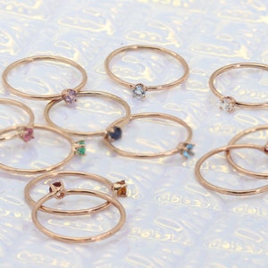 May include: A collection of delicate gold rings with colorful gemstones. Each ring features a single gemstone in a variety of colors, including blue, green, purple, and red.