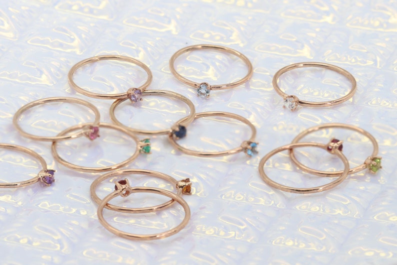 May include: A collection of delicate gold rings with colorful gemstones. Each ring features a single gemstone in a variety of colors, including blue, green, purple, and red.