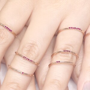 Puede incluir: Cinco anillos delicados de oro con una fila de peque&ntilde;as piedras preciosas rojas. Los anillos est&aacute;n apilados en los dedos de una persona.