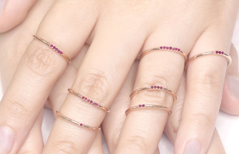 Puede incluir: Cinco anillos delicados de oro con una fila de peque&ntilde;as piedras preciosas rojas. Los anillos est&aacute;n apilados en los dedos de una persona.