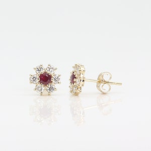 May include: A pair of gold earrings with a red gemstone surrounded by smaller white gemstones in a flower shape.