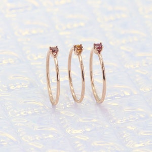 May include: Three delicate gold rings with small gemstones. The rings are stacked on a white background. The gemstones are red, orange, and yellow.