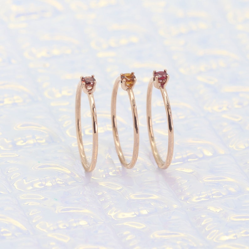 May include: Three delicate gold rings with small gemstones. The rings are stacked on a white background. The gemstones are red, orange, and yellow.