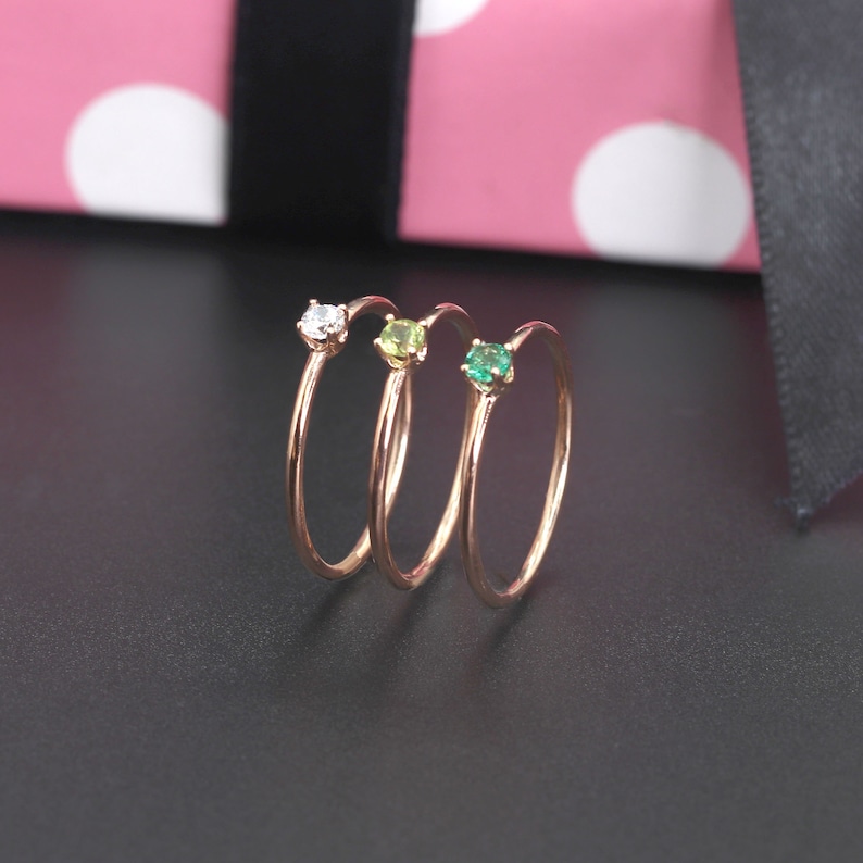 May include: Three delicate gold rings with small gemstones. The rings are stacked on top of each other. The gemstones are a clear diamond, a light green peridot, and a darker green emerald.