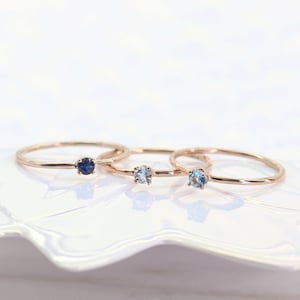 May include: Three delicate gold rings with small gemstones. The ring on the left has a dark blue gemstone, the middle ring has a light blue gemstone, and the ring on the right has a slightly darker blue gemstone.