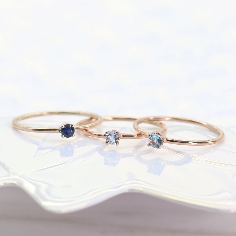May include: Three delicate gold rings with small gemstones. The ring on the left has a dark blue gemstone, the middle ring has a light blue gemstone, and the ring on the right has a slightly darker blue gemstone.