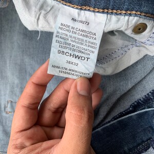 K&ouml;nnte beinhalten: Ein blaues Jeans-Etikett mit wei&szlig;em Text, der lautet: "RN130273 Made in Cambodia Hecho en Camboya 84% Cotton, 14% Polyester, 2% Spandex 84% Algodon, 14% Poli&eacute;ster, 2% Spandek Exclusive of Decoration/ Excepto Decoracion 98CHWDT 38X32 HHM-17K-MMMH26474 191056243975"