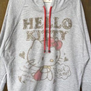 Op de afbeelding: Grijze hoodie met een Hello Kitty-print. De print toont Hello Kitty zittend met een rode strik en de tekst "How do you do I'm a white kin Look at my fa red Ruboo!" en "Look at me!"