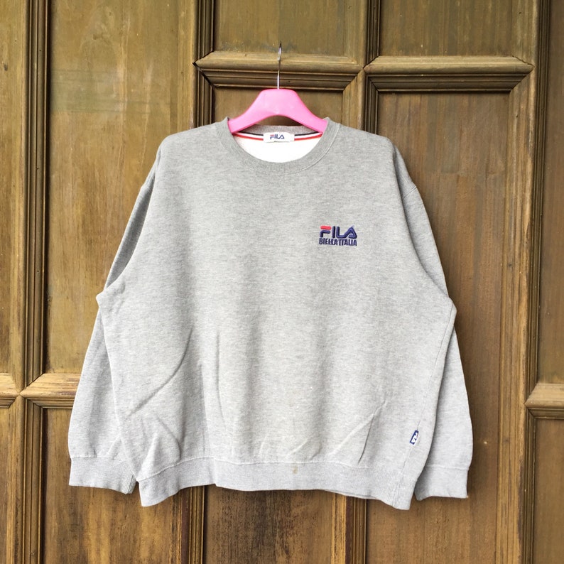 May include: Gray sweatshirt with a Fila logo embroidered on the front. The logo is in red, blue, and white and says "Fila BIELLA ITALIA".