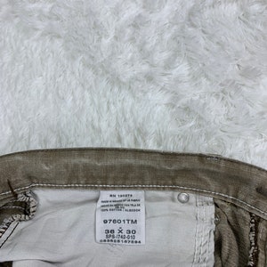 Op de afbeelding: Een wit label met zwarte tekst bevestigd aan een bruine spijkerbroek. Het label vermeldt "RN 130273 Made in Mexico of US Fabric Hecho en Mexico con Tela de 100% Cotton/Algodon 97601TM 36 X 30 SPS-1742-010 083625167594".