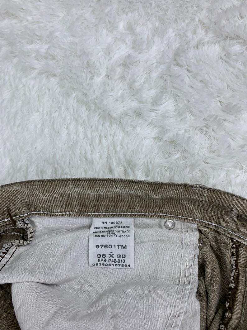 Op de afbeelding: Een wit label met zwarte tekst bevestigd aan een bruine spijkerbroek. Het label vermeldt "RN 130273 Made in Mexico of US Fabric Hecho en Mexico con Tela de 100% Cotton/Algodon 97601TM 36 X 30 SPS-1742-010 083625167594".