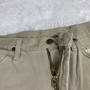 Op de afbeelding: Een beige werkbroek met een ritsvlieg en een knoopsluiting. De broek heeft een enkele voorzak en twee achterzakken.