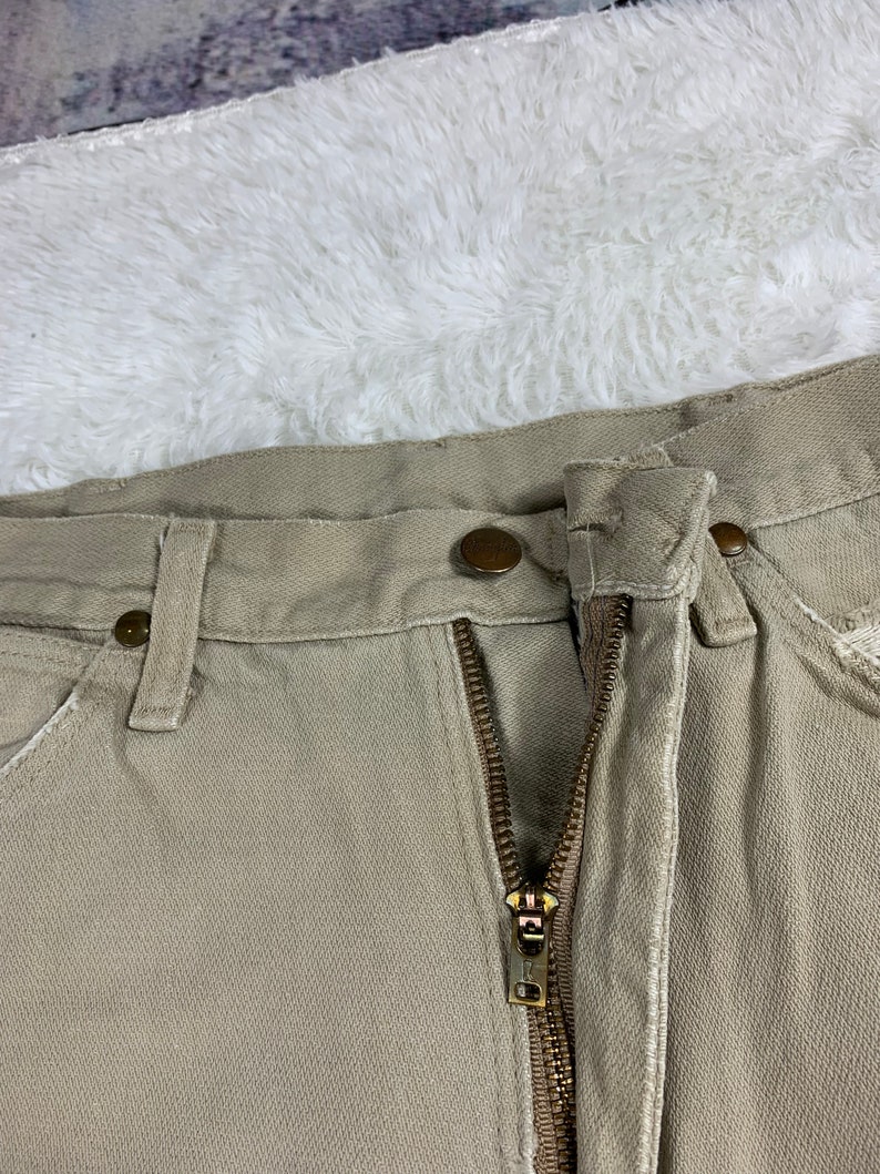 Op de afbeelding: Een beige werkbroek met een ritsvlieg en een knoopsluiting. De broek heeft een enkele voorzak en twee achterzakken.