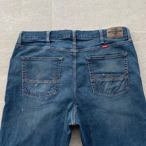 K&ouml;nnte beinhalten: Eine blaue Jeanshose mit einem braunen Leder-Patch auf der rechten Ges&auml;&szlig;tasche, auf dem "Wrangler Premium Valley" steht.
