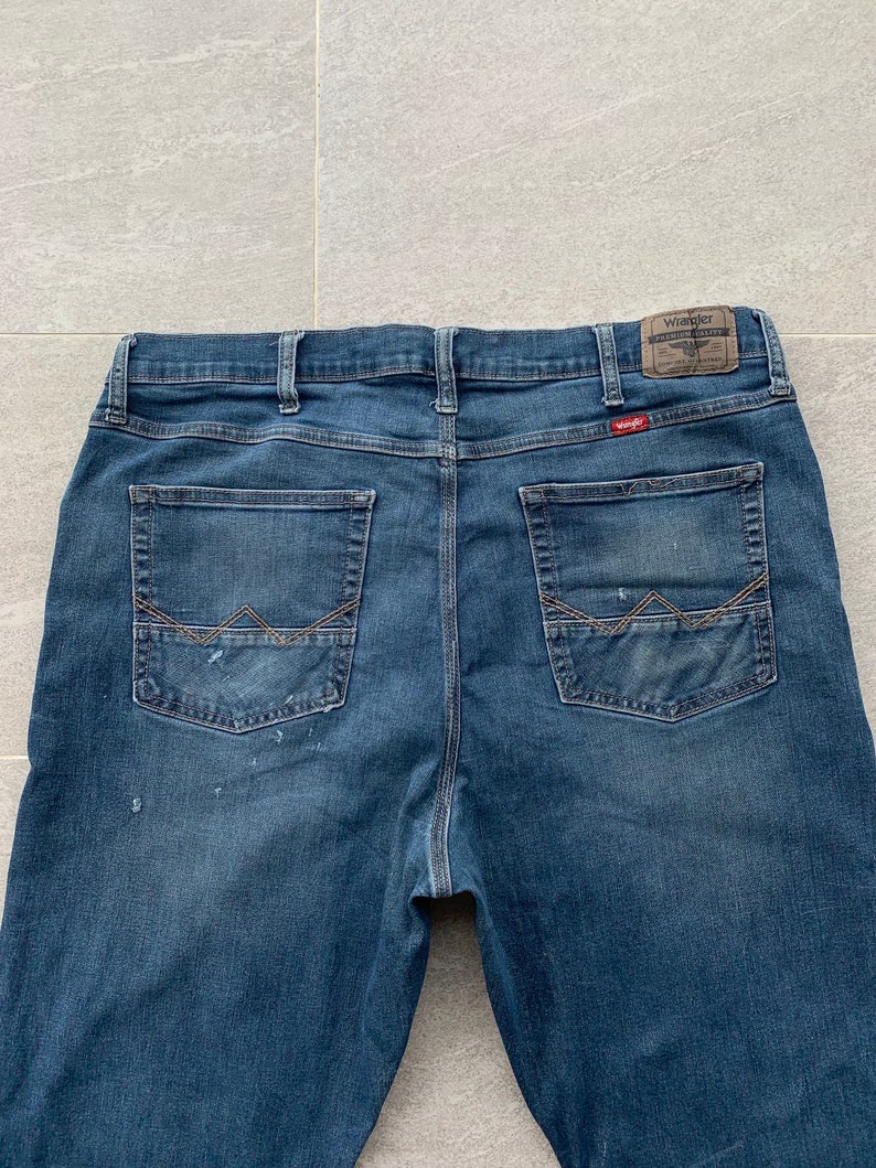 K&ouml;nnte beinhalten: Eine blaue Jeanshose mit einem braunen Leder-Patch auf der rechten Ges&auml;&szlig;tasche, auf dem "Wrangler Premium Valley" steht.