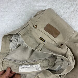Op de afbeelding: Een lichtbruine Wrangler jeans met een bruine leren patch op de achterzak. De jeans heeft een maatlabel met de tekst "Wrangler 936TAN 30 x 34" en "100% Katoen".