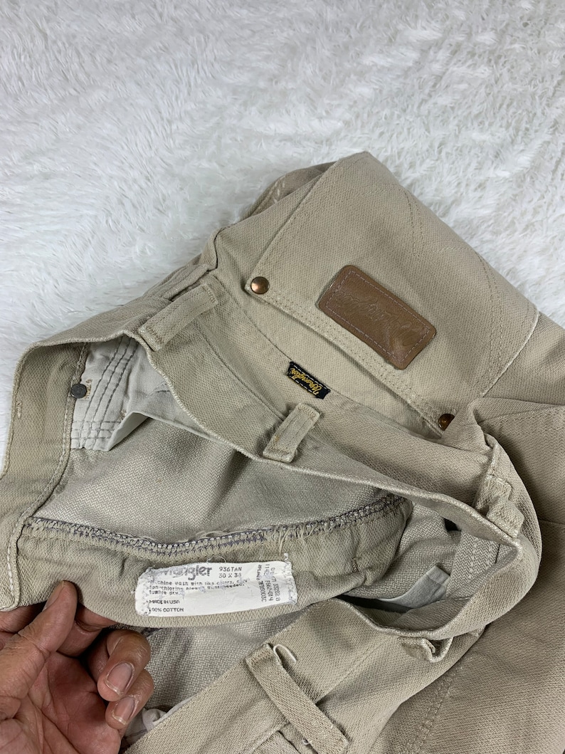 Op de afbeelding: Een lichtbruine Wrangler jeans met een bruine leren patch op de achterzak. De jeans heeft een maatlabel met de tekst "Wrangler 936TAN 30 x 34" en "100% Katoen".
