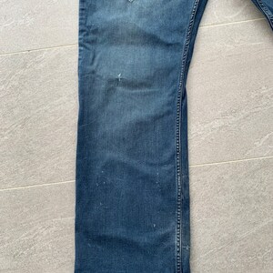 K&ouml;nnte beinhalten: Eine blaue Jeanshose mit Bootcut-Beinabschluss. Die Jeans hat einen Used-Look mit mehreren kleinen L&ouml;chern und verblassten Stellen.