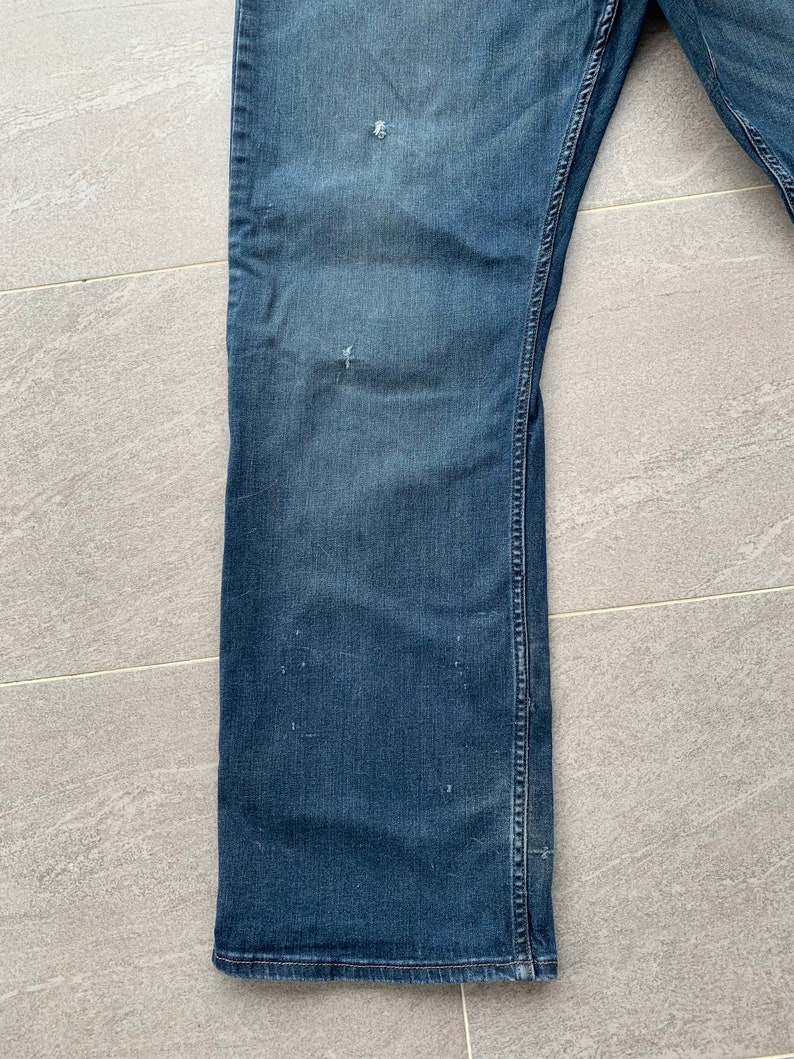 K&ouml;nnte beinhalten: Eine blaue Jeanshose mit Bootcut-Beinabschluss. Die Jeans hat einen Used-Look mit mehreren kleinen L&ouml;chern und verblassten Stellen.