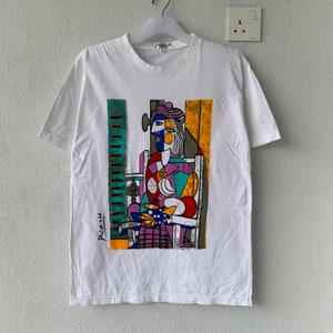 Puede incluir: Camiseta blanca con un estampado gráfico colorido de una mujer sentada en una silla. El gráfico es una pintura de estilo cubista de Picasso. La camiseta tiene cuello redondo y mangas cortas.