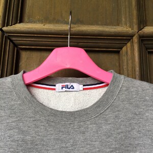 May include: Gray sweatshirt with a white Fila logo on the front. The sweatshirt has a ribbed neckline and a white, red, and blue stripe around the neckline.