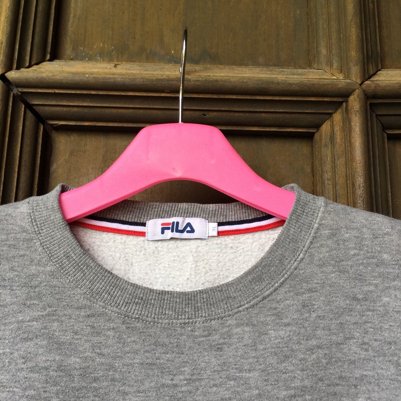 May include: Gray sweatshirt with a white Fila logo on the front. The sweatshirt has a ribbed neckline and a white, red, and blue stripe around the neckline.