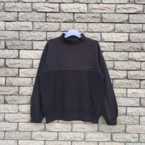 Könnte beinhalten: Ein dunkelbraunes Sweatshirt mit Stehkragen und einem kontrastierenden Band über der Brust. Das Sweatshirt hat das Wort "kouvr" auf dem Band gestickt.