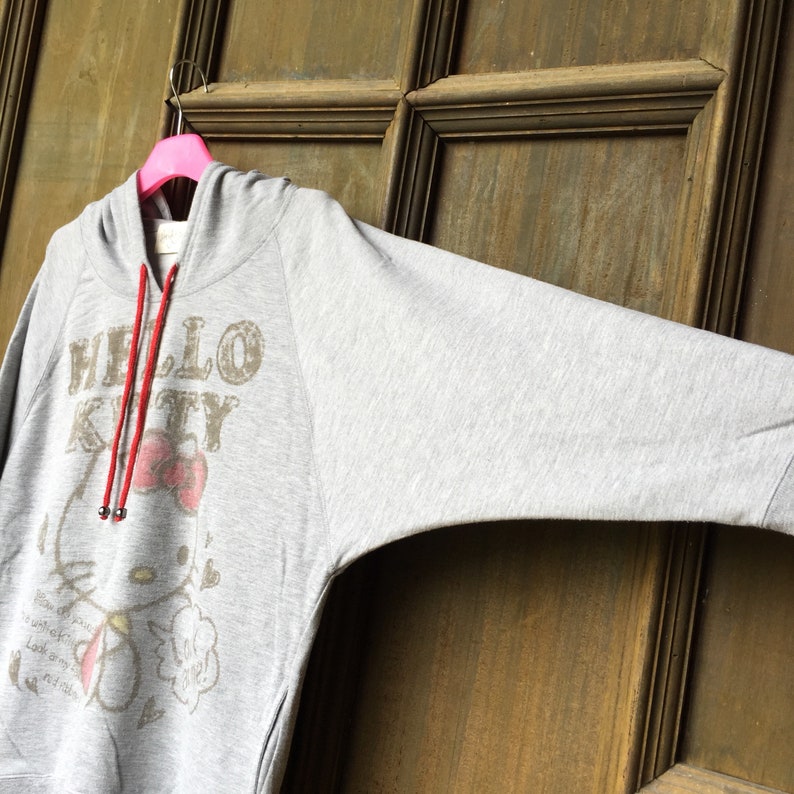 Op de afbeelding: Een grijs hoodie met een Hello Kitty-print en een rood trekkoord. De print toont Hello Kitty met een rode strik en de woorden "Hello Kitty" in een verweerd lettertype.